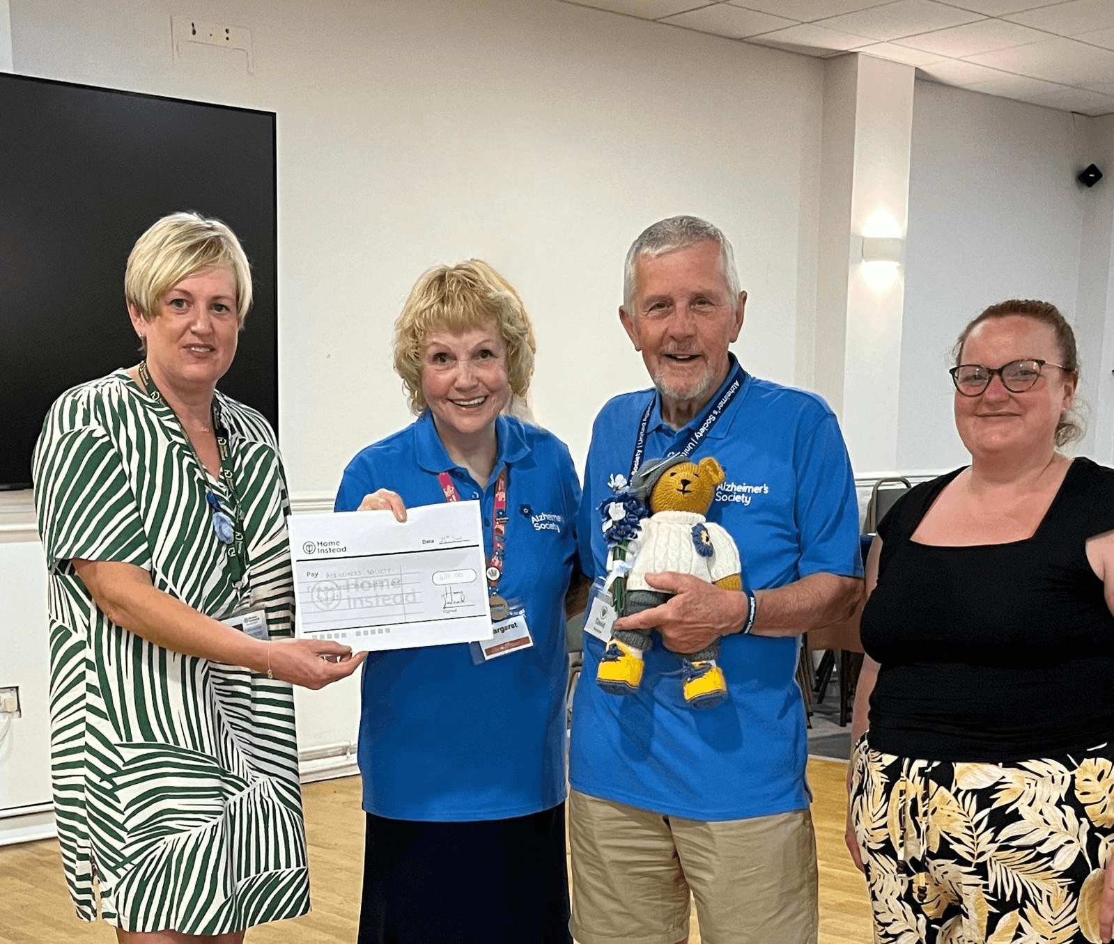 Four adults stand indoors; one holds a cheque, another a teddy bear, all smiling at the camera. - Home Instead