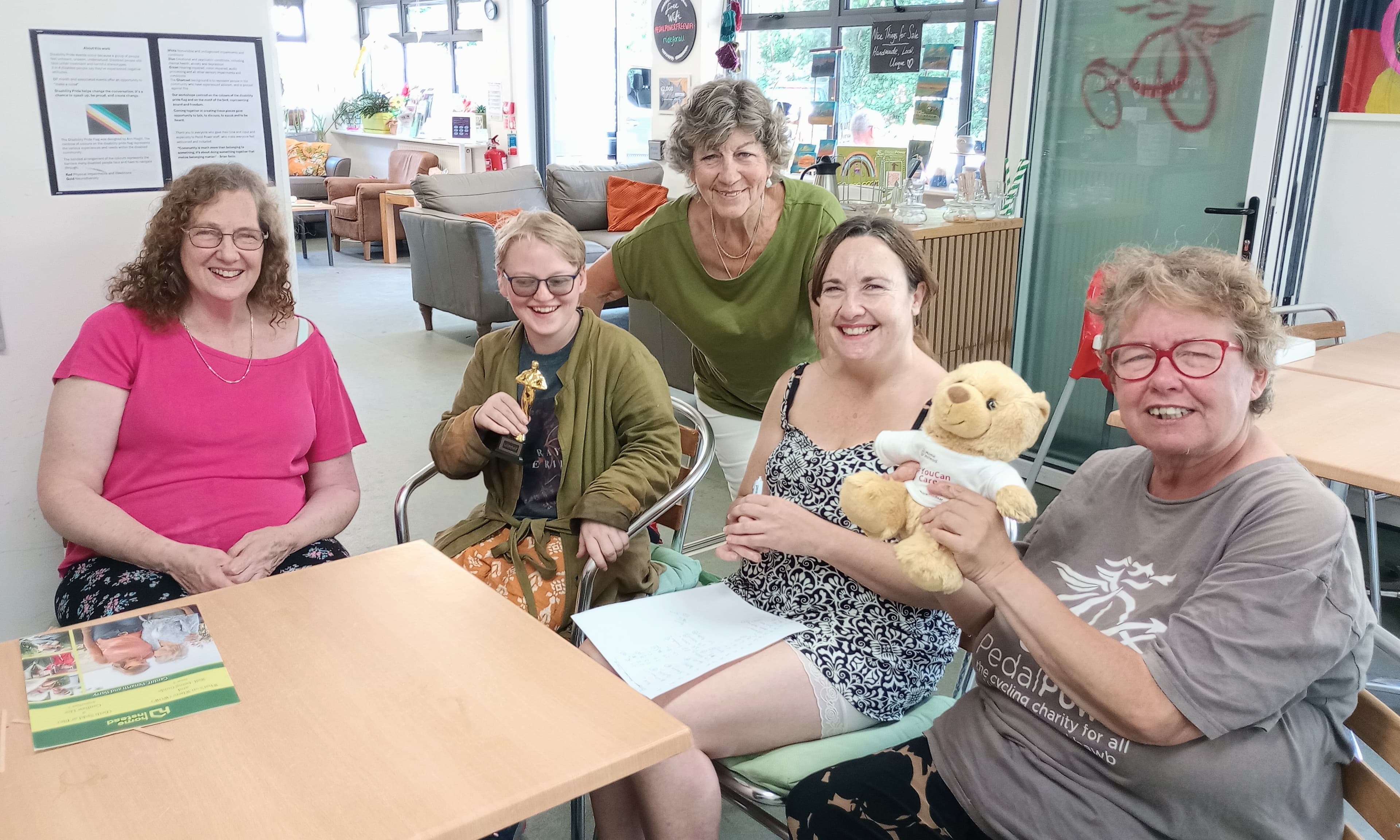 Five smiling women sit together indoors, one holding a teddy bear, in a casual and cheerful setting. - Home Instead