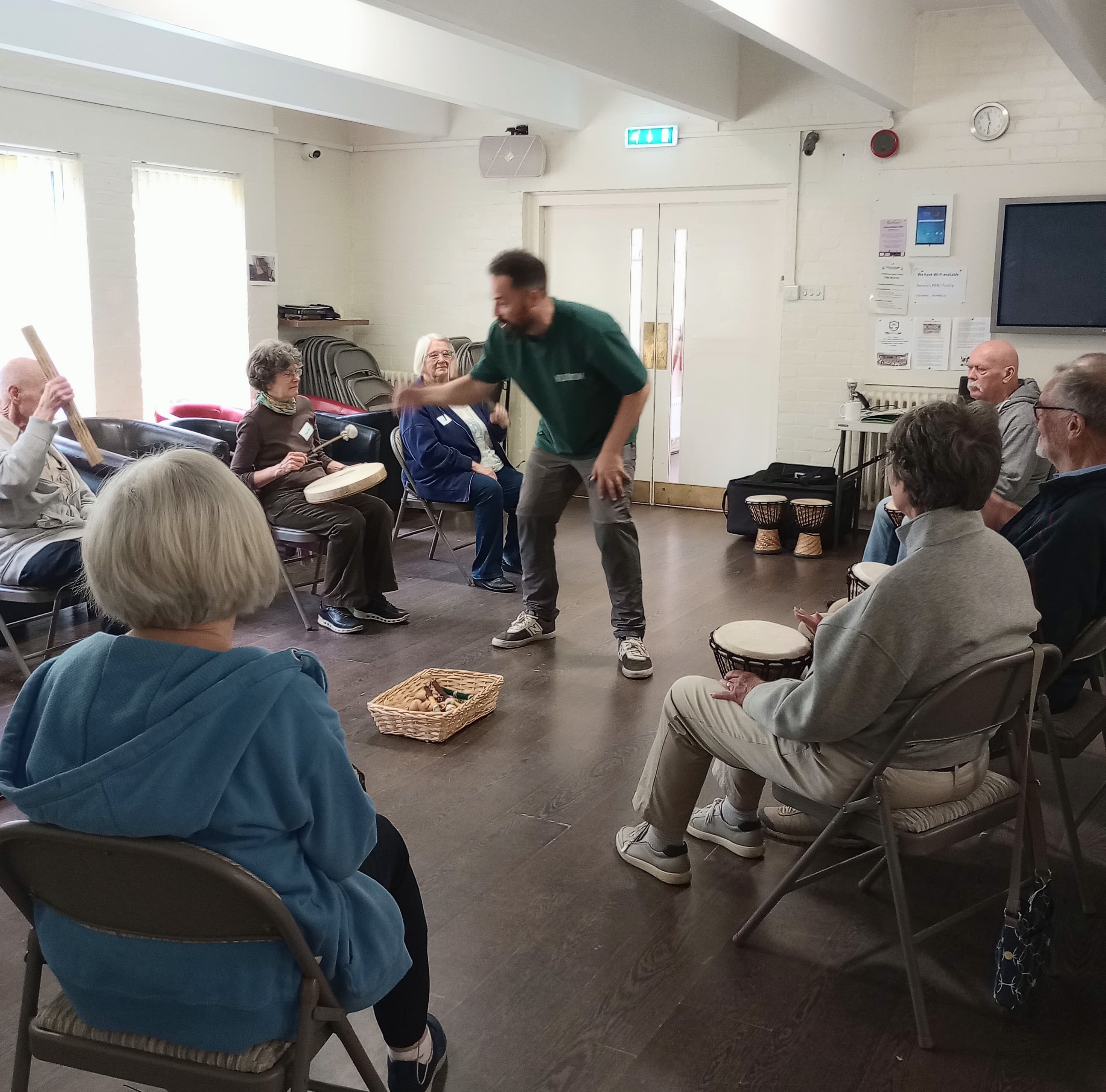 A group of older adults sit in a circle playing drums and percussion with an instructor standing in the middle. - Home Instead