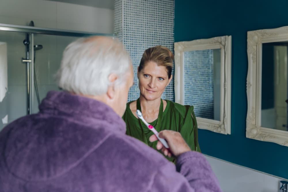 An elderly man holds a toothbrush whilst a woman in green stands beside him in a bathroom. - Home Instead