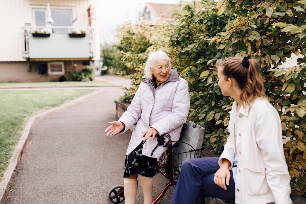 Older woman on a Zimmer frame smiles and talks with a young woman outside by green bushes on a pavement. - Home Instead