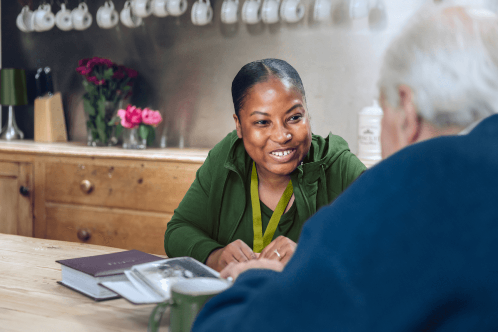 A woman smiling and talking with an older man at a wooden table with books and flowers nearby. - Home Instead