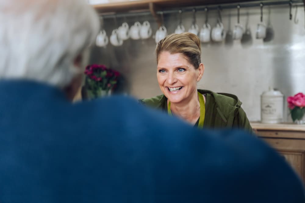 Smiling woman with blonde hair talks to an older man in a cosy kitchen with mugs hanging behind her. - Home Instead