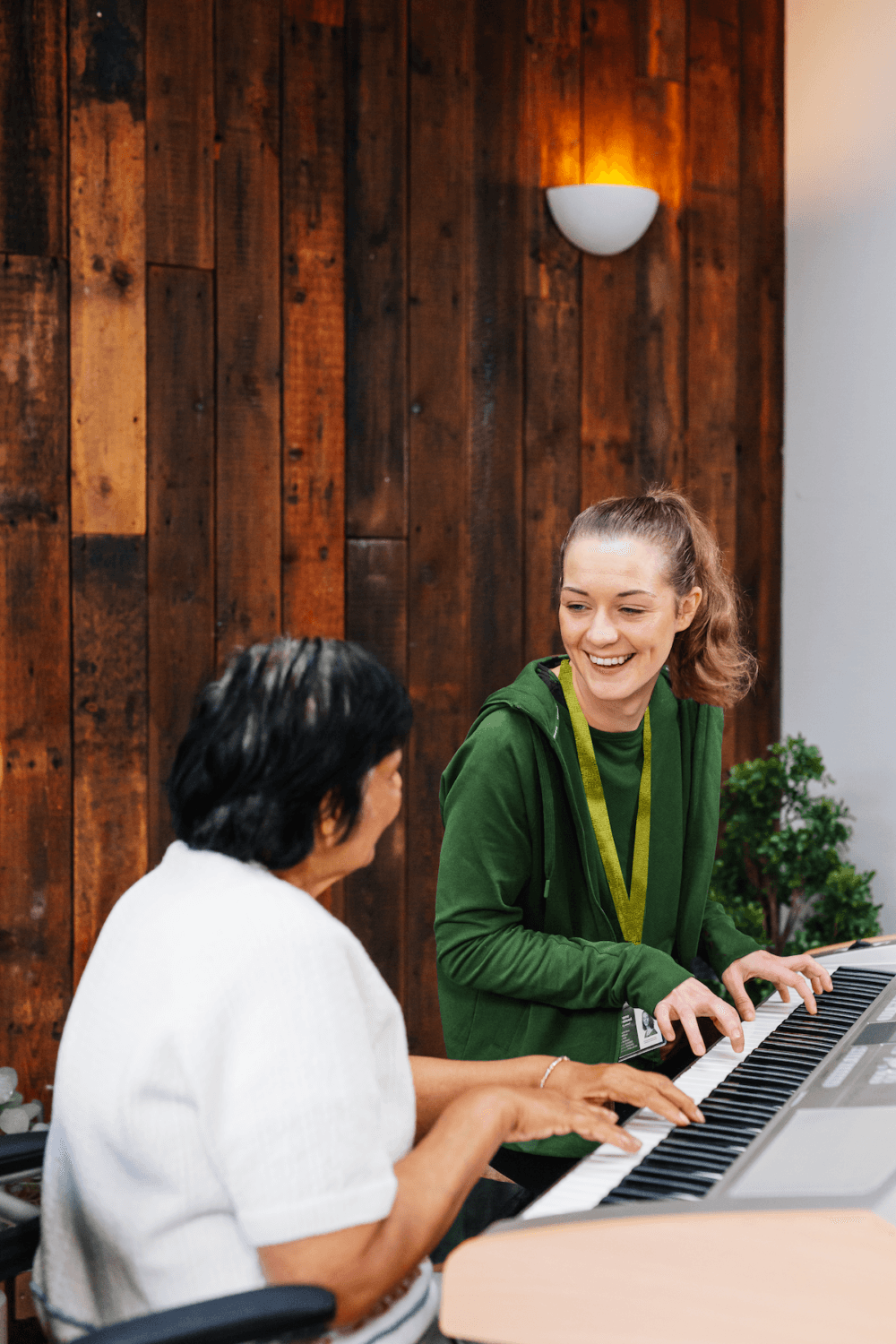 Two women smiling and playing an electric keyboard together in a cosy room with wooden walls. - Home Instead