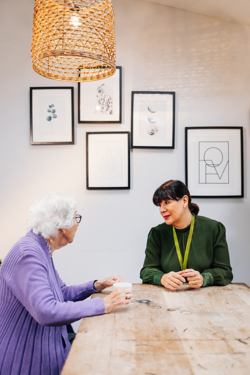 An elderly woman and a younger woman sit at a wooden table, talking under a wicker lampshade. - Home Instead