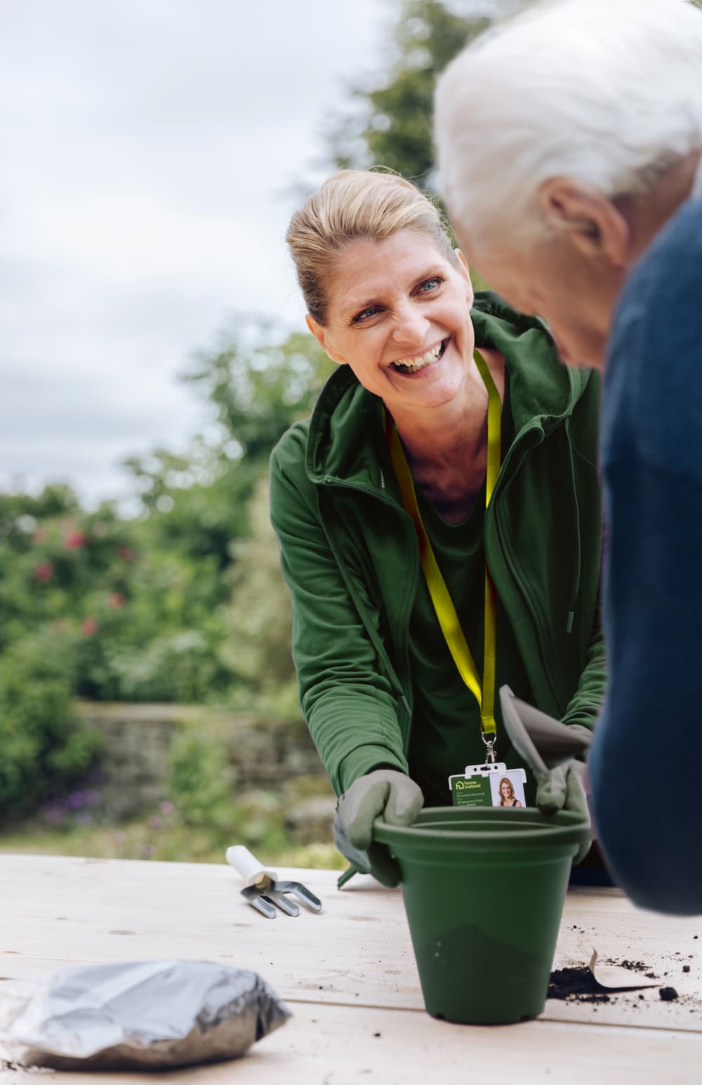 Smiling woman in green helps an elderly person with gardening, holding a green plant pot outdoors. - Home Instead