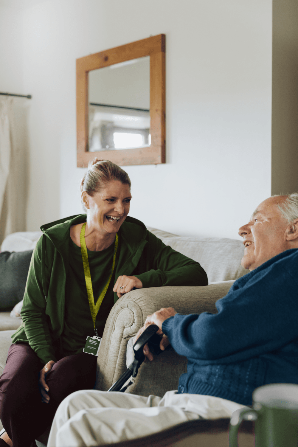 A woman in a green hoodie smiles whilst talking to an elderly man sitting on a sofa with a walking stick. - Home Instead