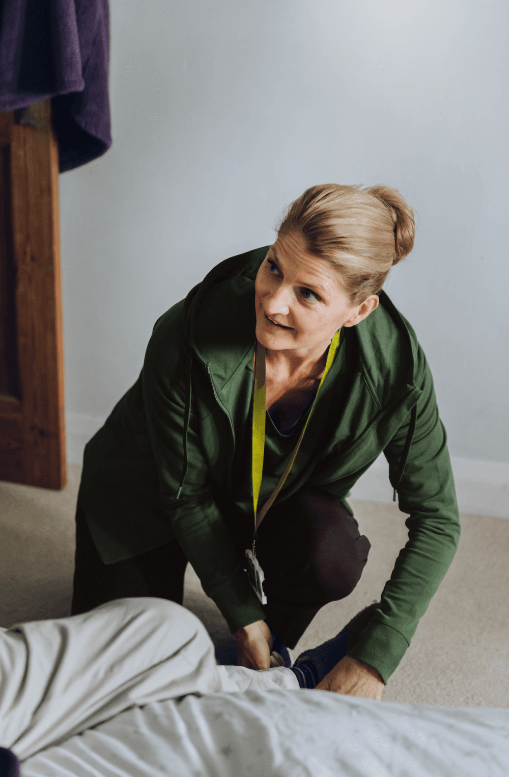 A woman in a green jacket kneels by a bed, helping someone put on socks. - Home Instead