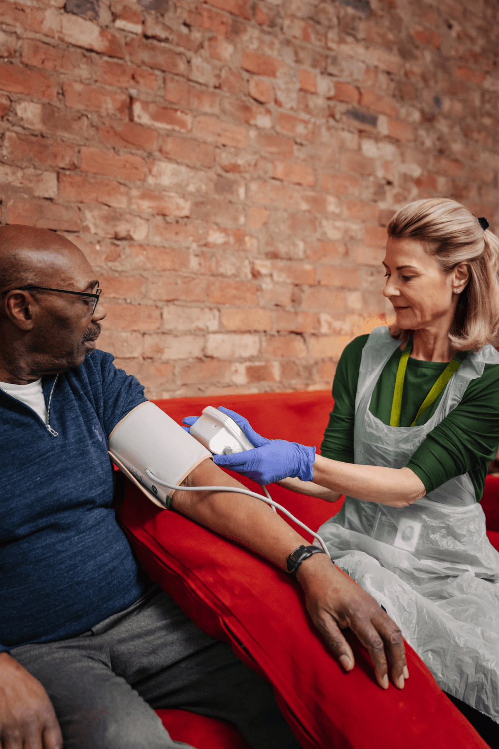 A nurse checks a man's blood pressure as they sit on a red sofa against a brick wall. - Home Instead