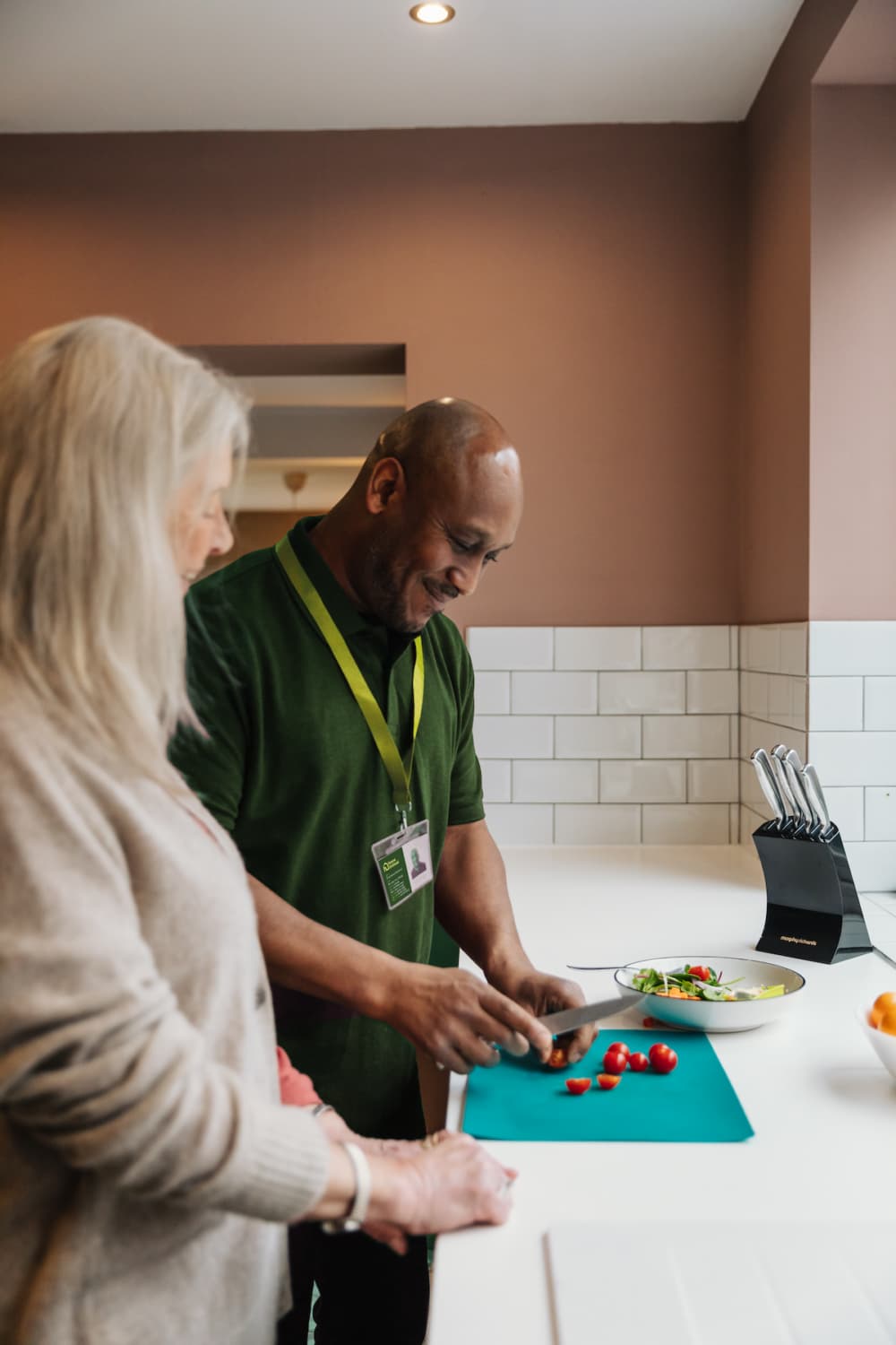 A man cuts tomatoes on a board while a woman watches in a kitchen with white tiles. - Home Instead