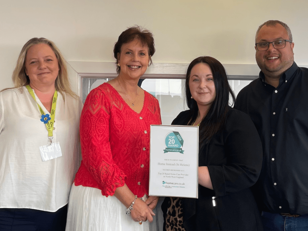 Four people smiling, one holding a framed award certificate, standing together indoors near a window. - Home Instead