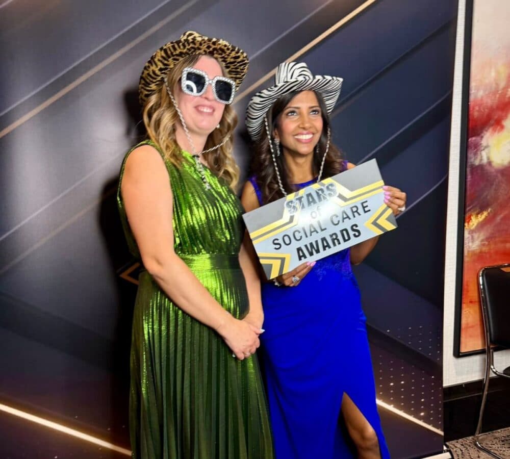 Two women in fancy hats and dresses pose, holding a "Stars of Social Care Awards" sign and smiling. - Home Instead