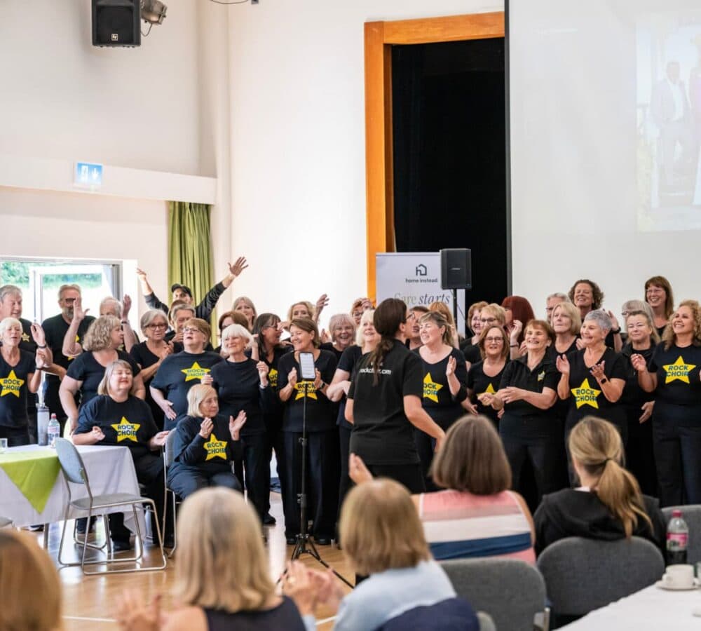 A choir of older adults performs on stage in matching shirts, while an audience applauds in a hall. - Home Instead