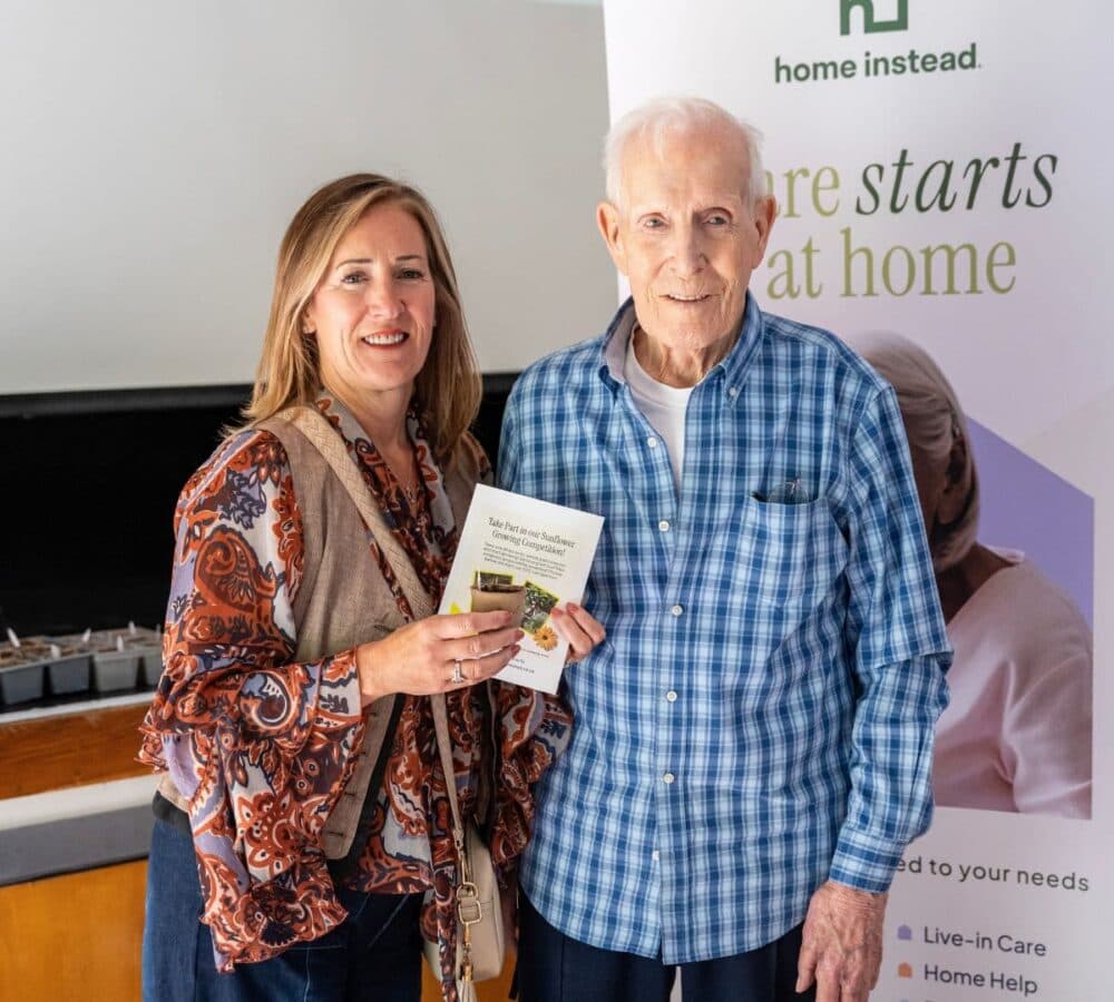A woman and an older man smile together, holding a booklet in front of a "Home Instead" care banner. - Home Instead