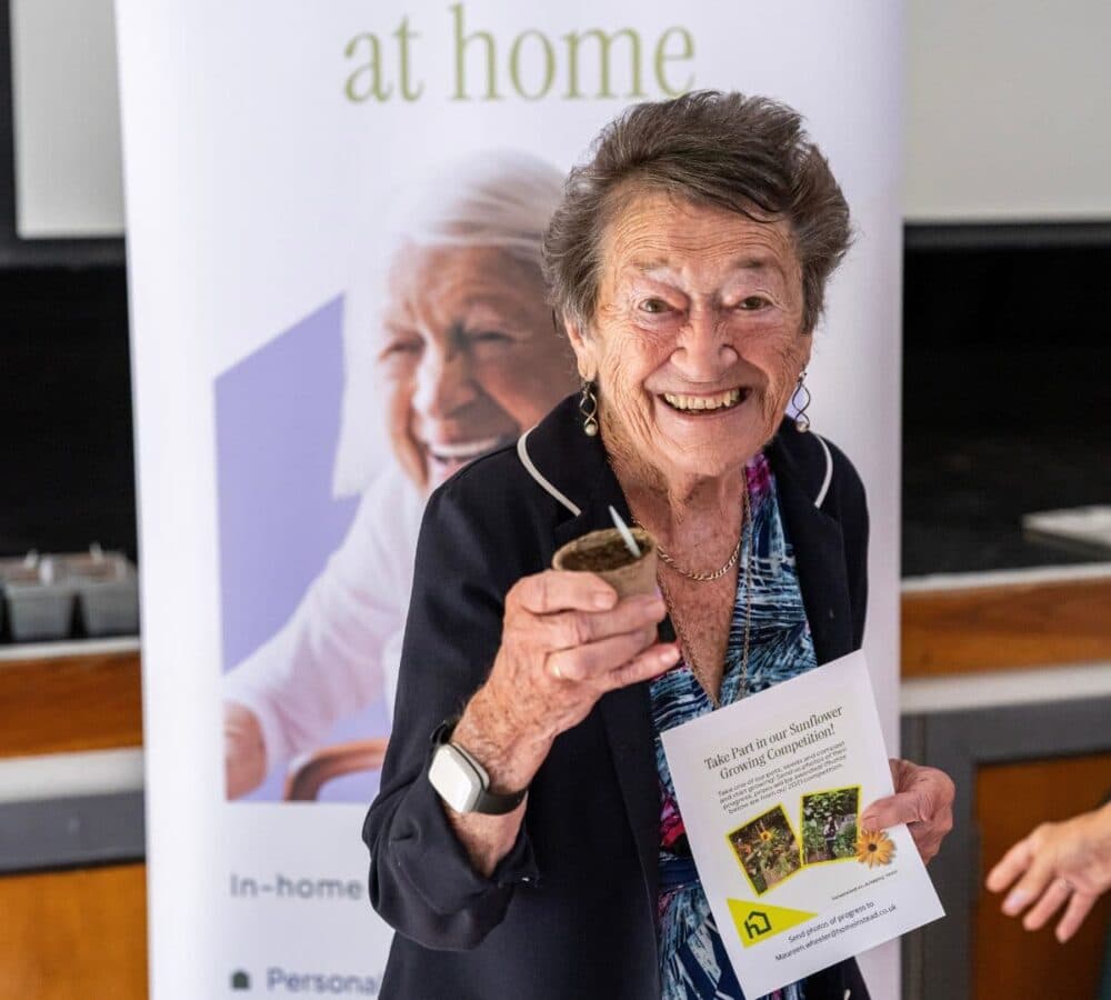 Smiling elderly woman holds a small plant and a leaflet, standing in front of a promotional banner. - Home Instead
