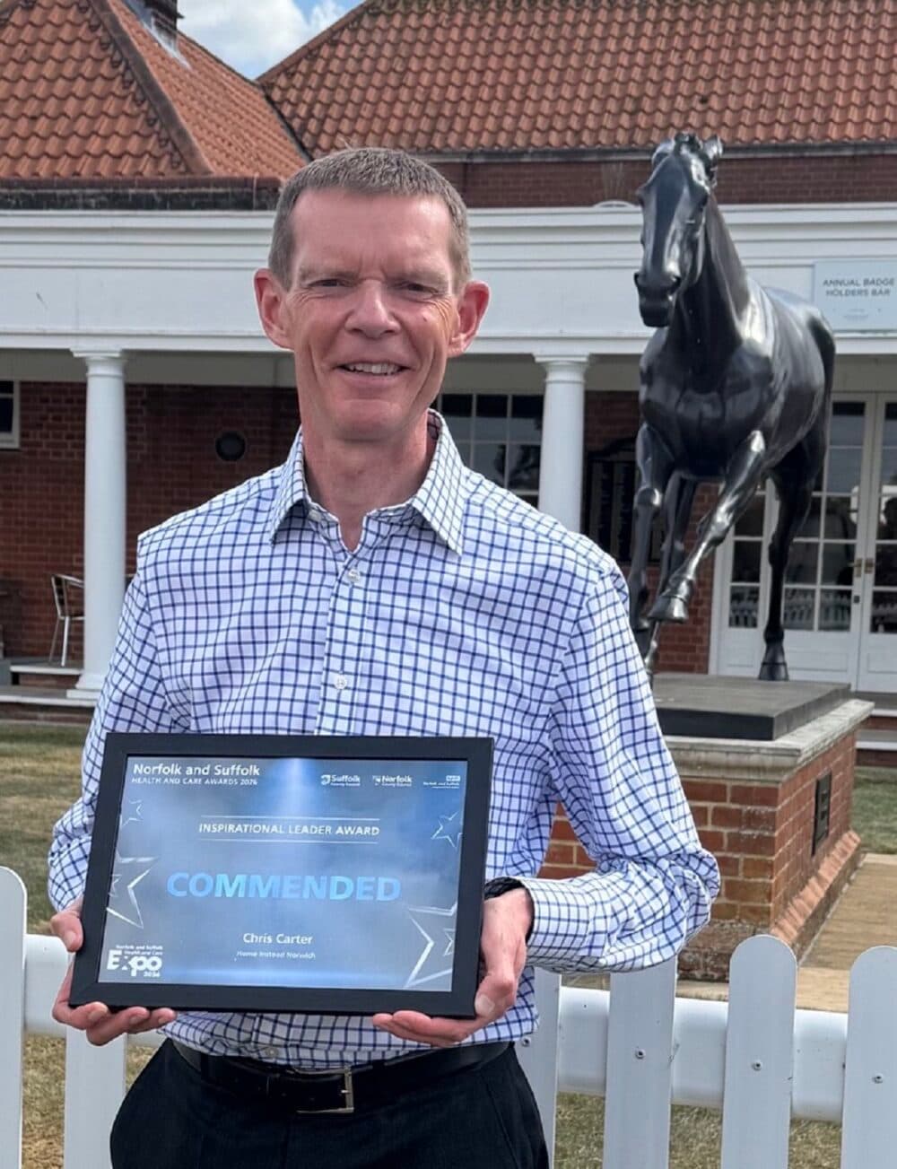 Man holding an award plaque, standing outside near a statue of a horse and a building with a red-tiled roof. - Home Instead