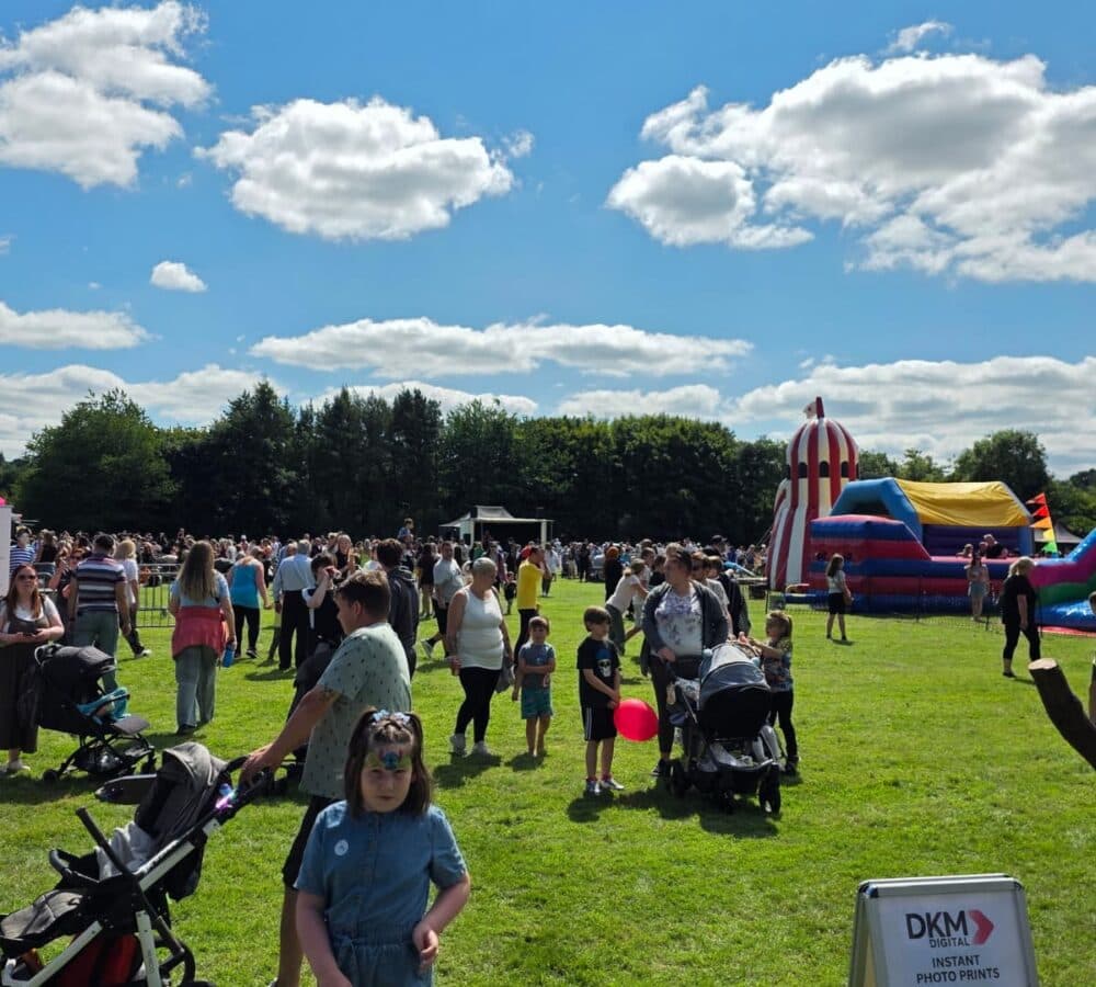 Crowd at an outdoor fair with bouncy castles, children, pushchairs, and a photo booth sign on a sunny day. - Home Instead