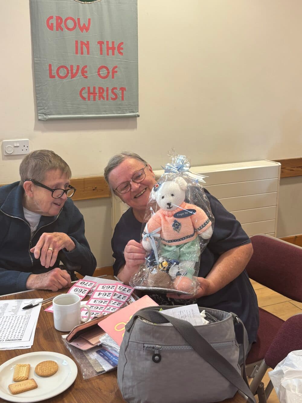 Two smiling women sit at a table with raffle tickets; one holds a teddy bear gift hamper. - Home Instead
