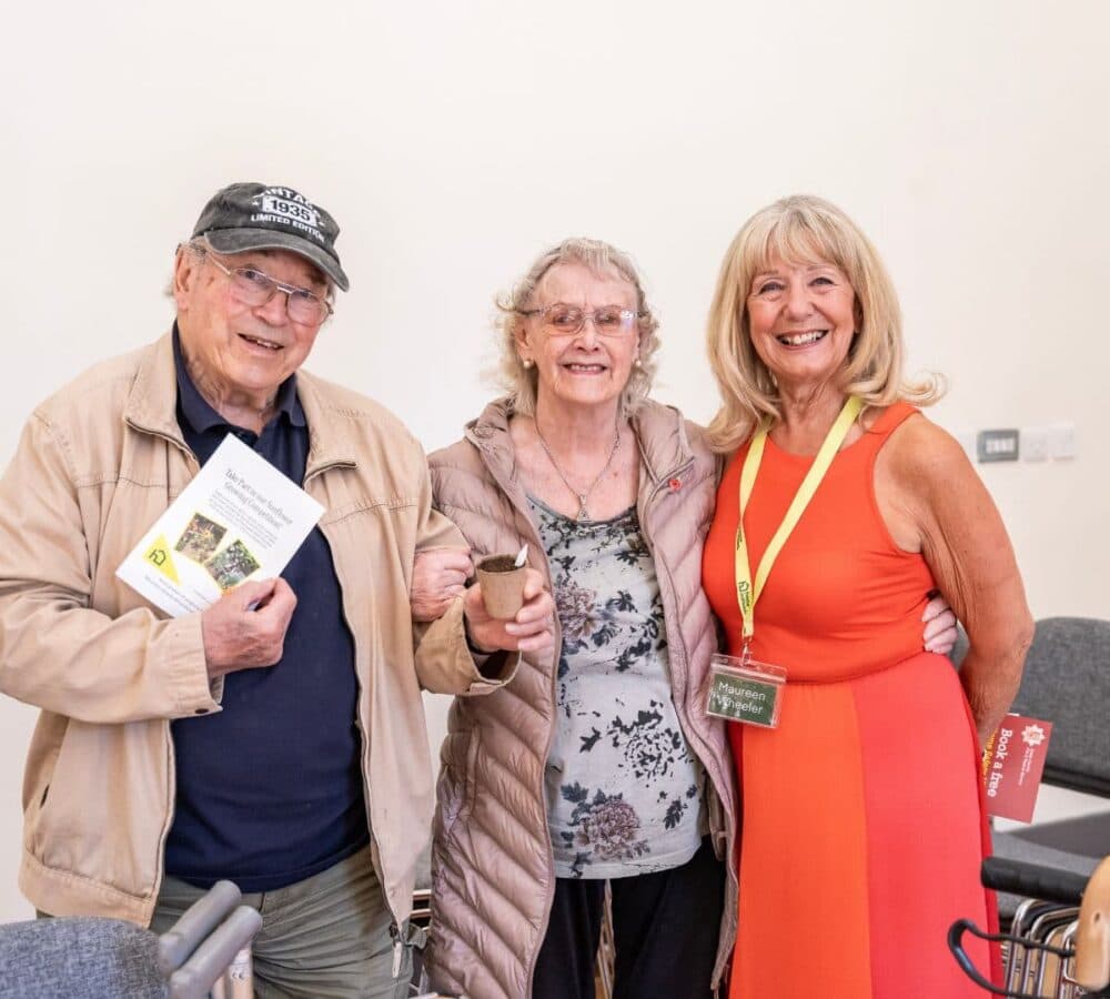 Three smiling older adults pose together indoors; one holds a booklet, another a cup, and one wears a name badge. - Home Instead