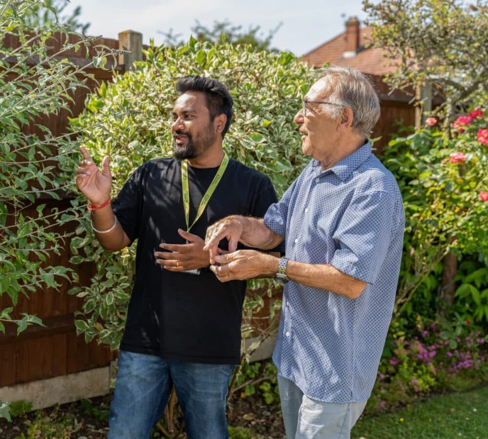 Two men standing outside in a garden, talking and smiling on a sunny day. - Home Instead