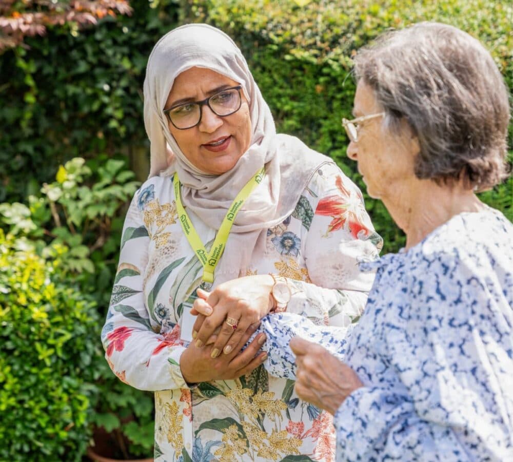 A woman in a headscarf assists an older woman outdoors in a green garden, both smiling. - Home Instead