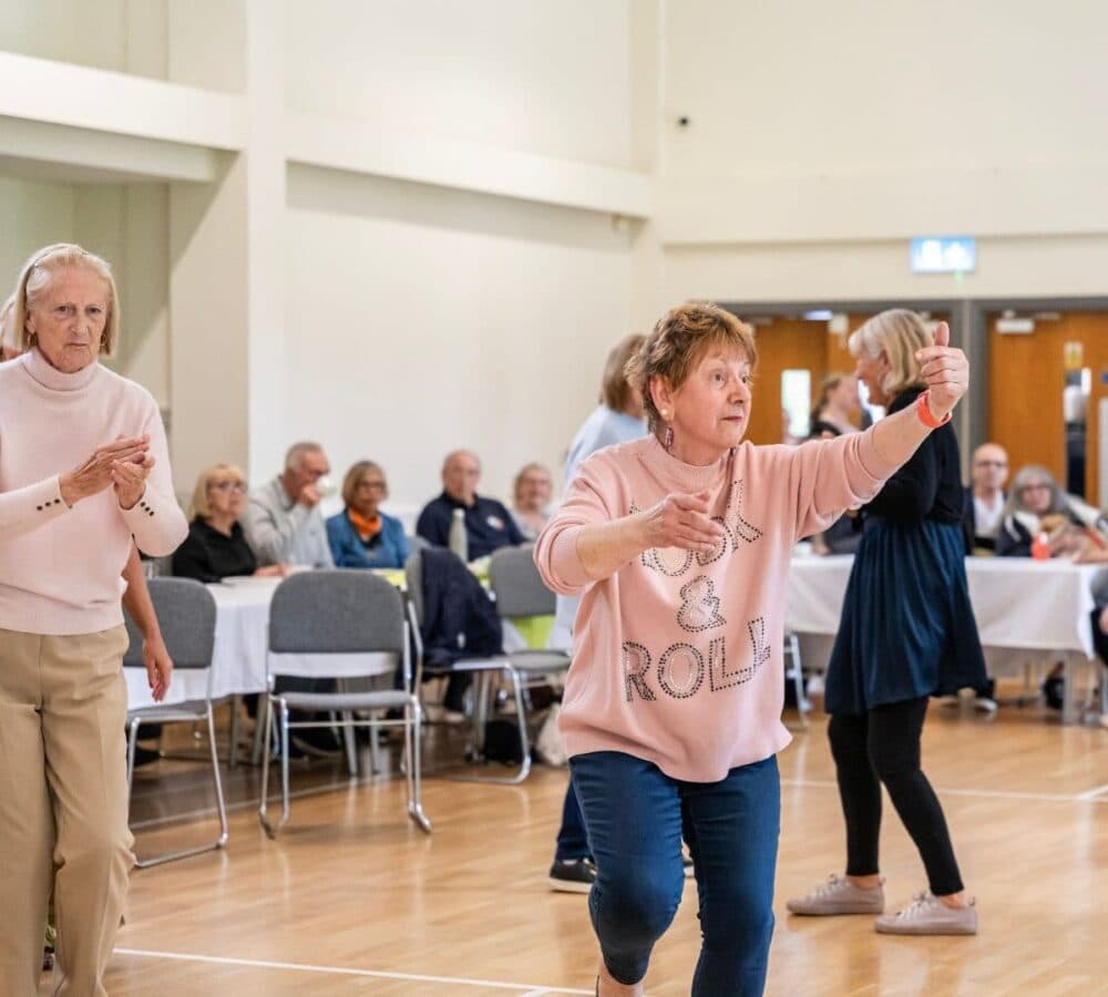 Older adults dancing energetically in a bright room with seated people watching in the background. - Home Instead