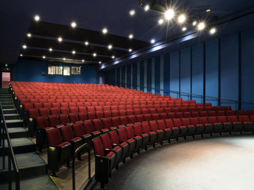 An empty auditorium with rows of red seats and stage lights on, facing towards a stage with a blue wall backdrop. - Home Instead