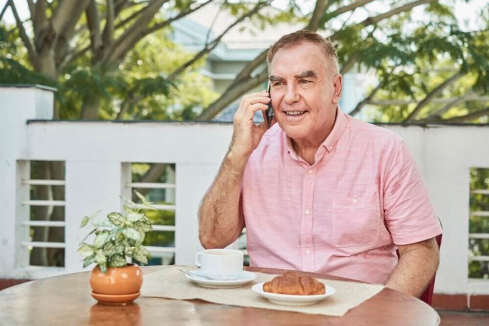 Smiling older man in a pink shirt talking on the phone while sitting at an outdoor table with coffee and a croissant. - Home Instead
