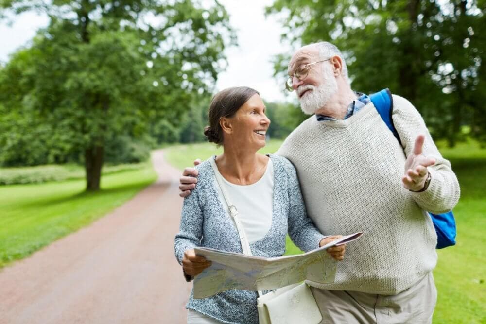 A senior couple smiles at each other while holding a map, walking on a path in a green park on a sunny day. - Home Instead Southampton