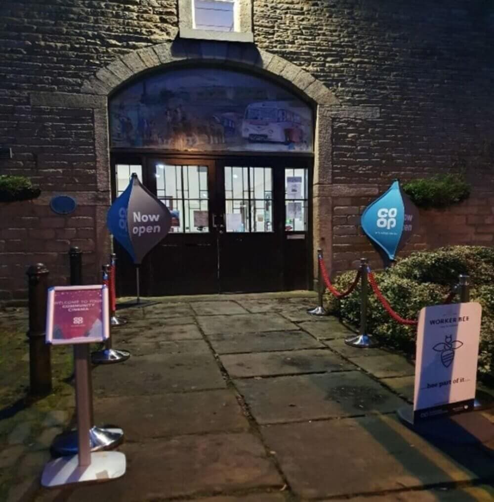 Entrance to a Co-op store with "Now open" signs and promotional displays on each side of the doorway. - Home Instead