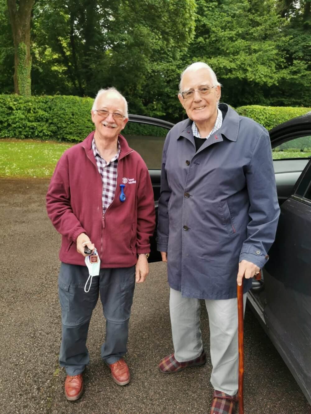 Two elderly men standing outside next to a car. One holds a device and the other has a walking stick. Greenery in the background. - Home Instead