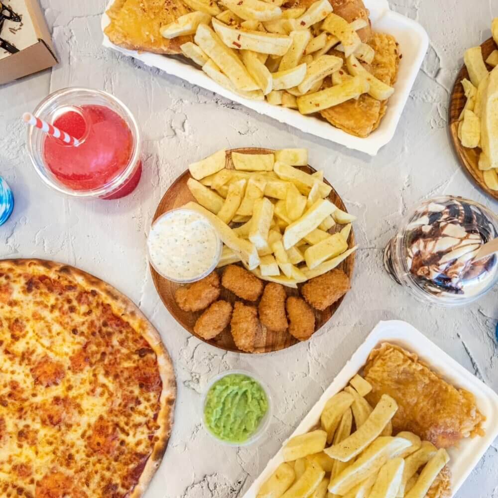 Andy's Golden Fry - A table filled with pizza, fish and chips, chicken nuggets, fries, guacamole, a pink drink, and a chocolate dessert. - Home Instead Renfrewshire and Barrhead home care services