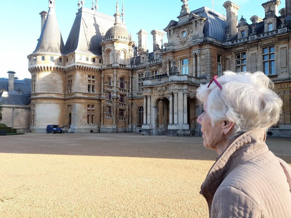 An elderly woman with white hair stands outside, gazing at a large, ornate historic building. - Home Instead