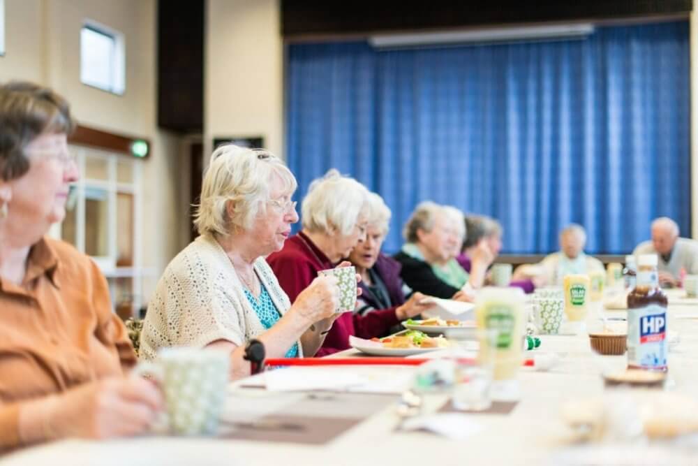 Elderly people sitting at a table, eating and drinking, with blue curtains in the background. - Home Instead