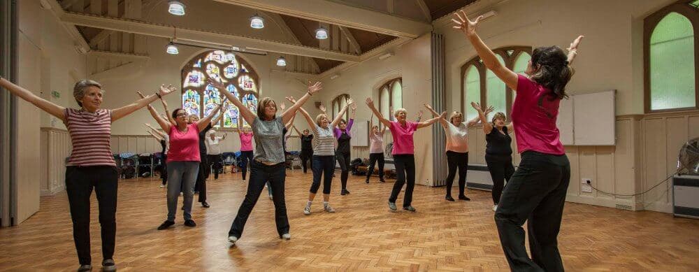 A group of people, led by an instructor, practice dance or exercise in a spacious hall with stained glass windows. - Home Instead