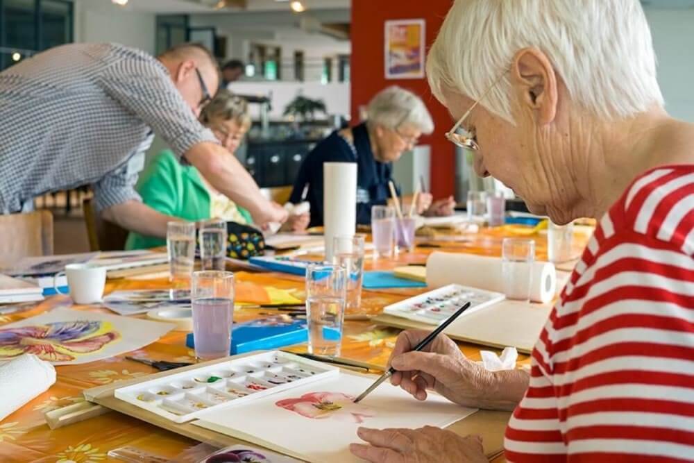 Seniors engaged in a watercolor painting class, focusing on their artwork at a table with art supplies. - Home Instead