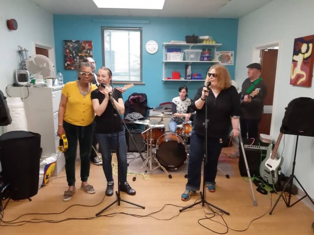 Band with two vocalists, guitarist, bassist, drummer, and tambourine player rehearsing in a colorful room with instruments. - Home Instead