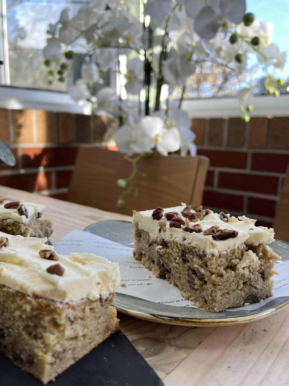 Two slices of frosted nut cake on plates with a brick wall and white flowers in the background. - Home Instead