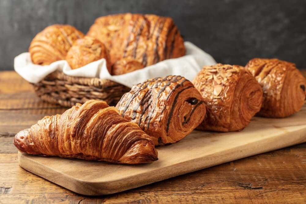 Assorted pastries, including croissants and pain au chocolat, on a wooden board and in a basket on a rustic table. - Home Instead