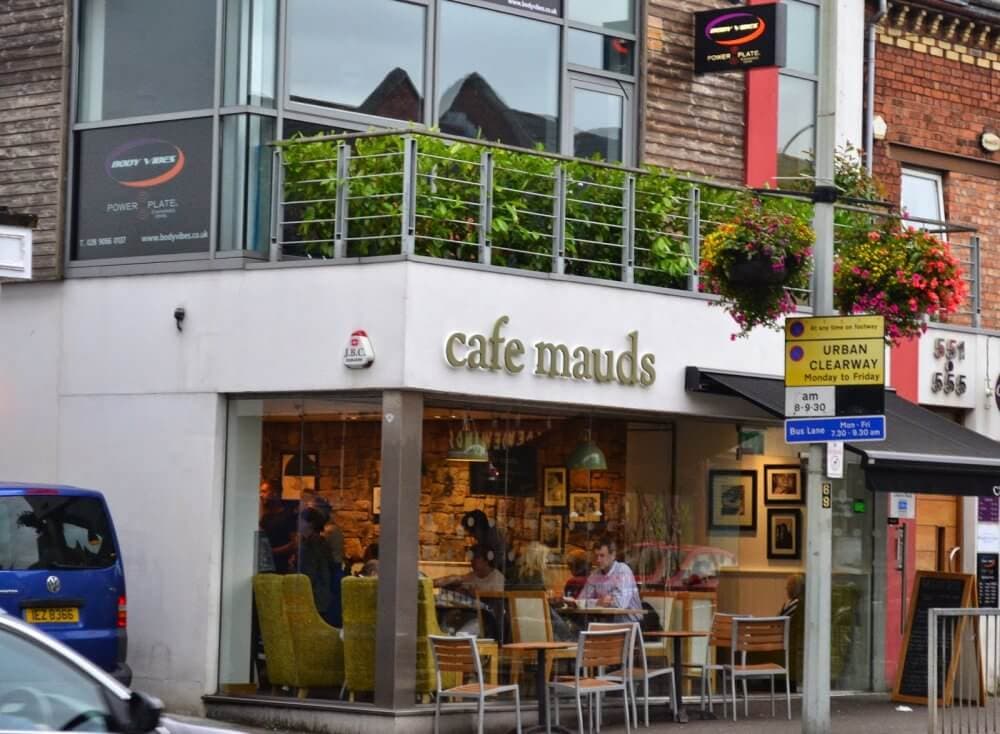 A street view of a café with the sign "cafe mauds," featuring outdoor seating and a hanging flower basket above. - Home Instead