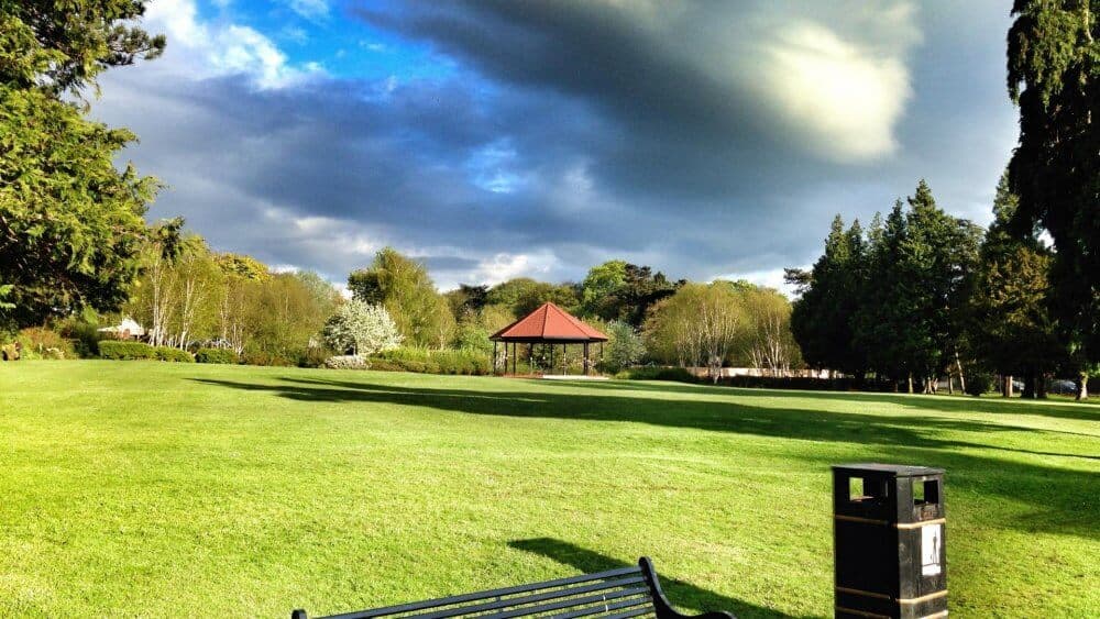 A park with a green lawn, a gazebo in the distance, a bench in the foreground, and trees under a cloudy sky. - Home Instead