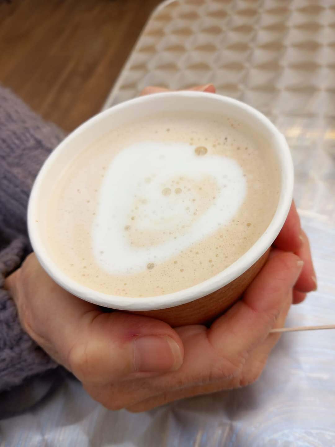 A person holding a cup of coffee with heart-shaped foam on top, over a patterned table. - Home Instead