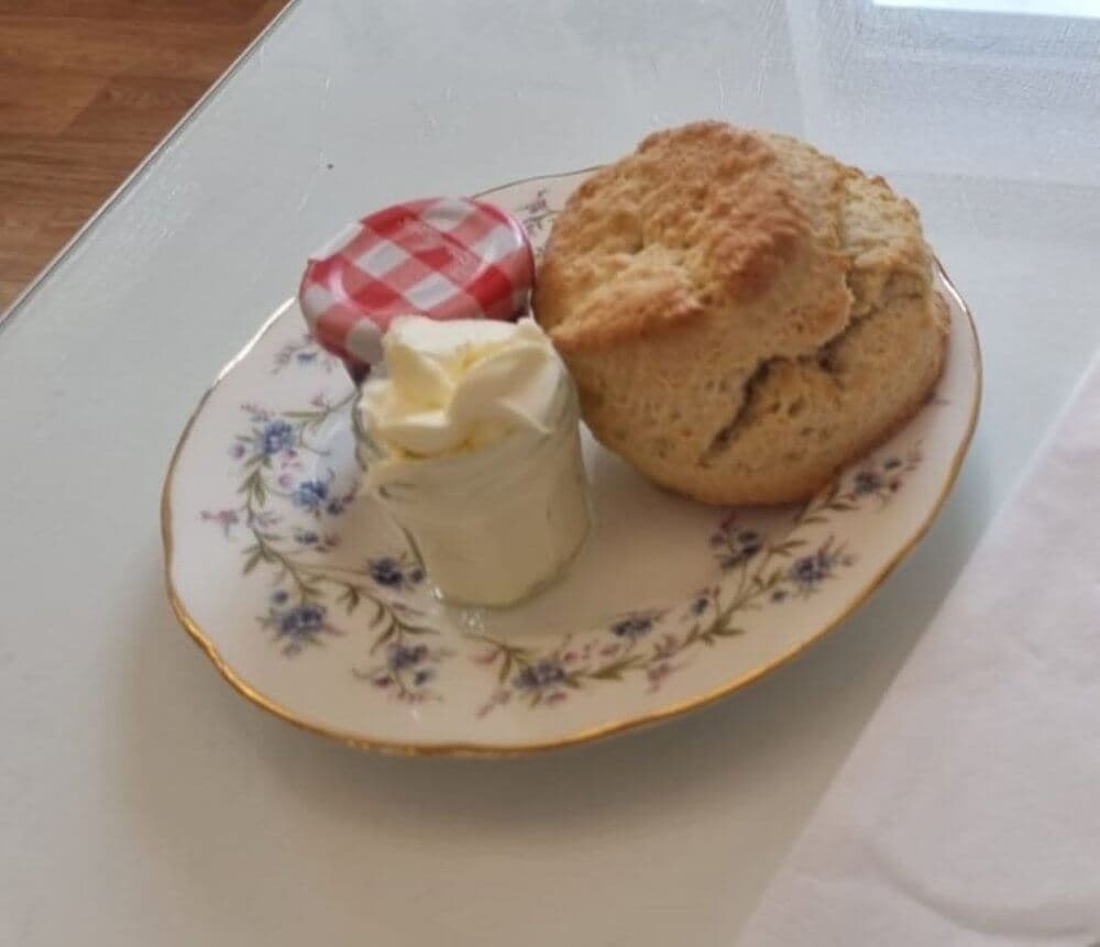 A scone sits on a floral plate next to cream in a small jar and a mini jar of strawberry jam with a red-checkered lid. - Home Instead