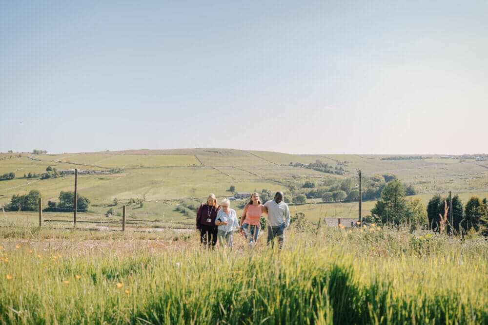 Four people walking through a grassy field under a clear blue sky, with rolling hills in the background. - Home Instead