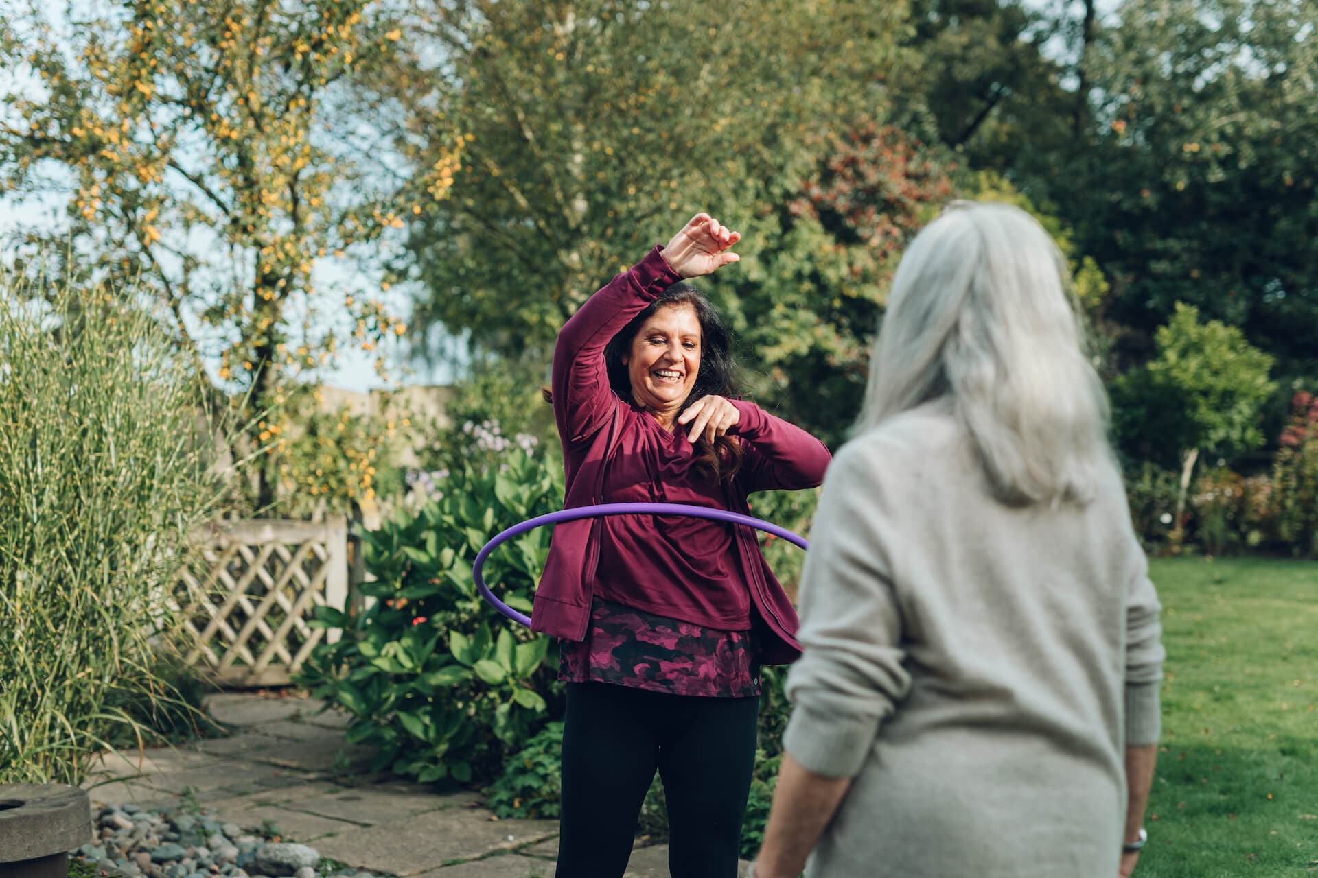 Two women enjoying time together outdoors, one hula-hooping and the other watching with trees in the background. - Home Instead