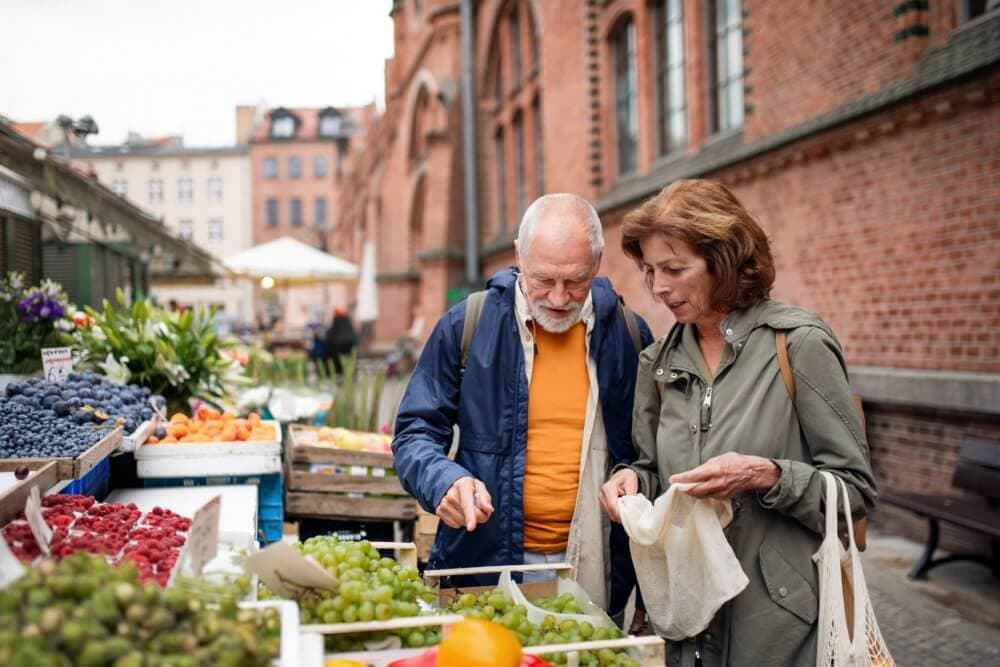An elderly couple browsing fresh produce at an outdoor market, with stalls of fruits and flowers around them. - Home Instead