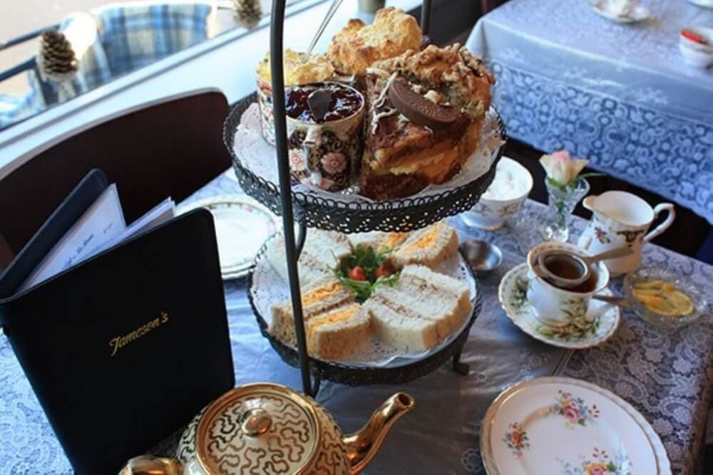 A tiered tray with scones, sandwiches, and desserts on a lace tablecloth, surrounded by teacups and a teapot. - Home Instead