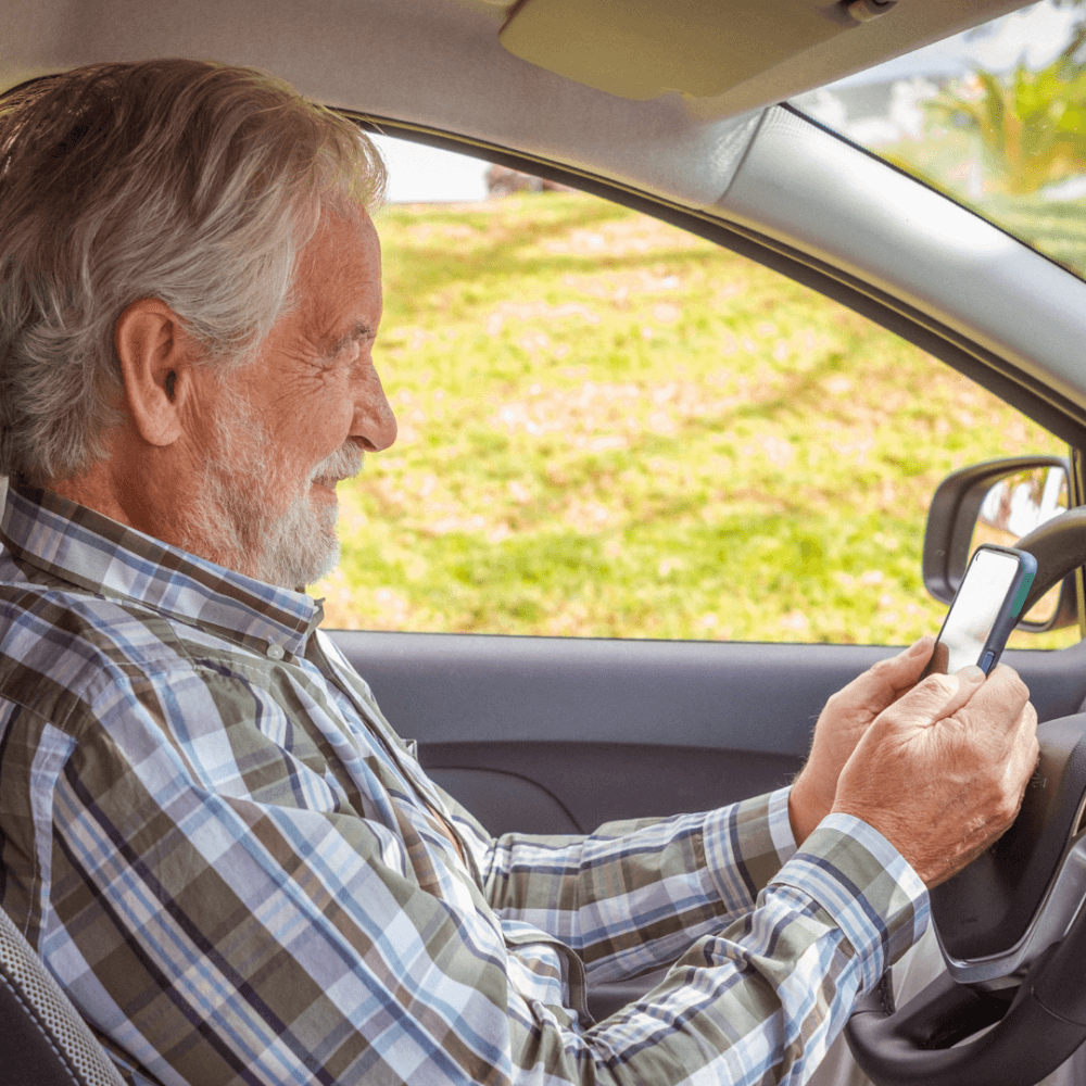Older man with gray hair and beard in a car smiling while looking at his smartphone. - Home Instead