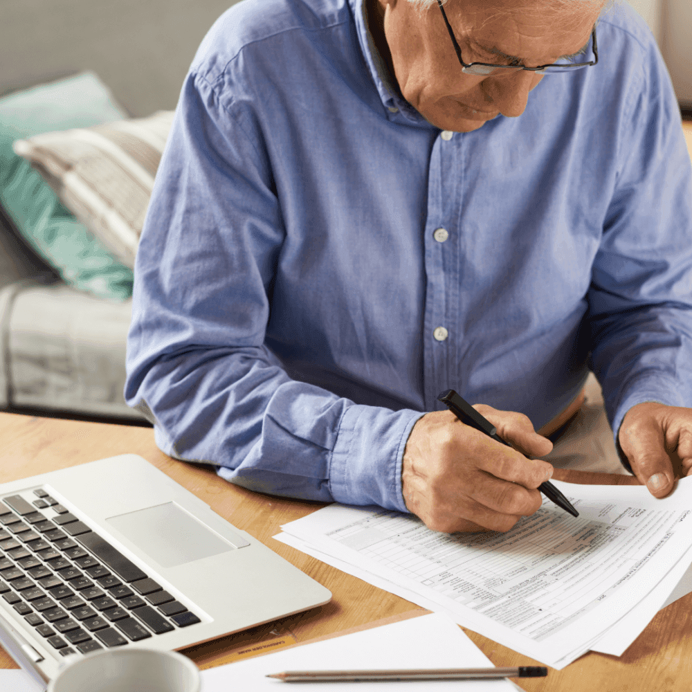 Older man in blue shirt writing on documents at a desk with a laptop and pen. - Home Instead