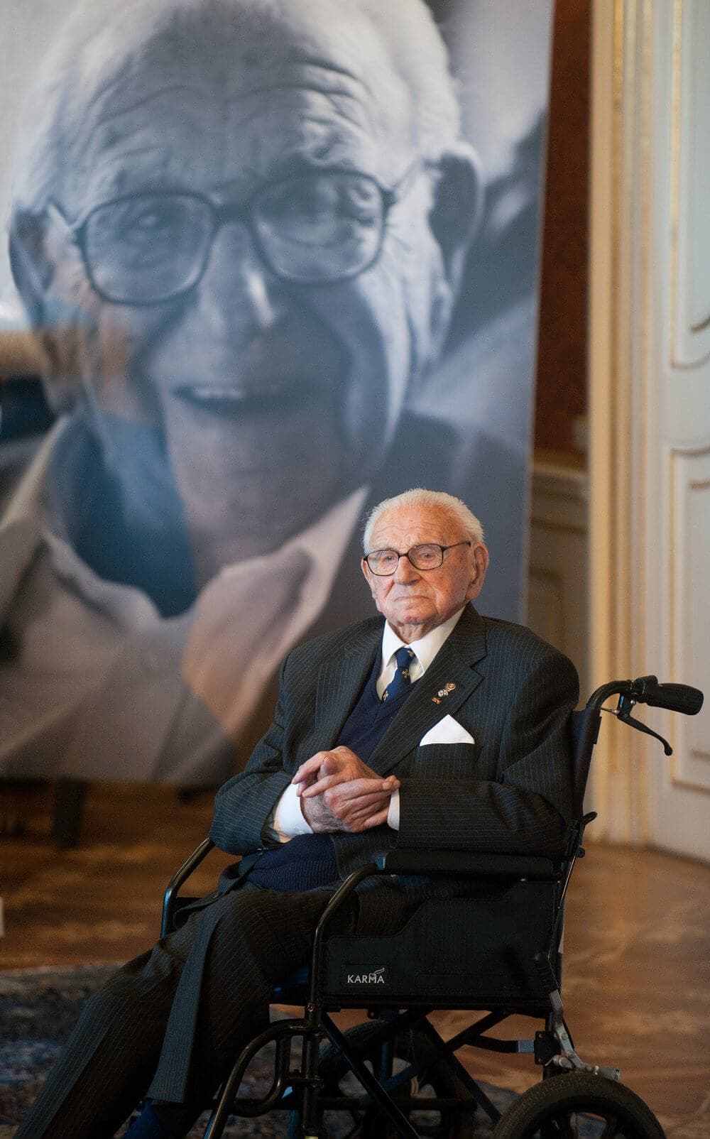An elderly man in a wheelchair wearing a suit sits in front of a large portrait of himself. - Home Instead